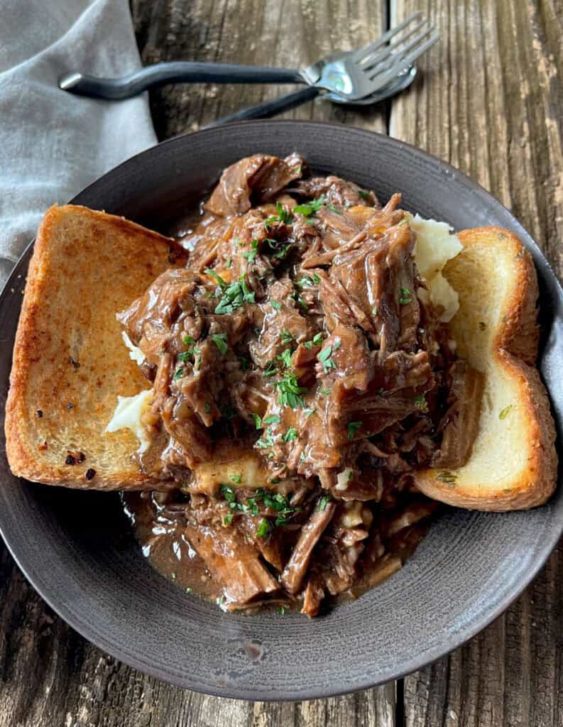 Hearty shredded beef and gravy served over mashed potatoes with toasted bread in a brown bowl on a rustic wooden table.