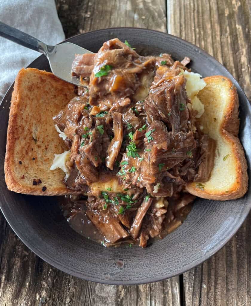 Hearty shredded beef and gravy served over mashed potatoes with toasted bread in a brown bowl on a rustic wooden table.