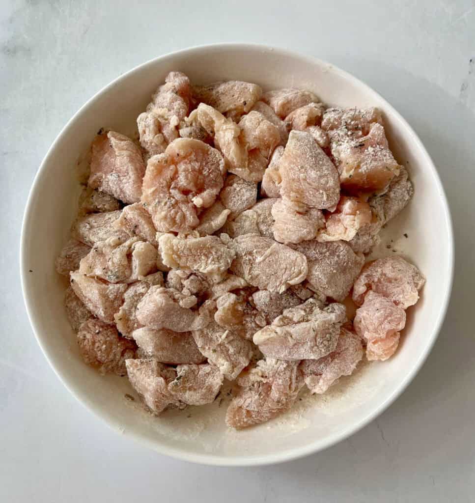 Lightly flour dusted chicken pieces in a white bowl on a white surface.