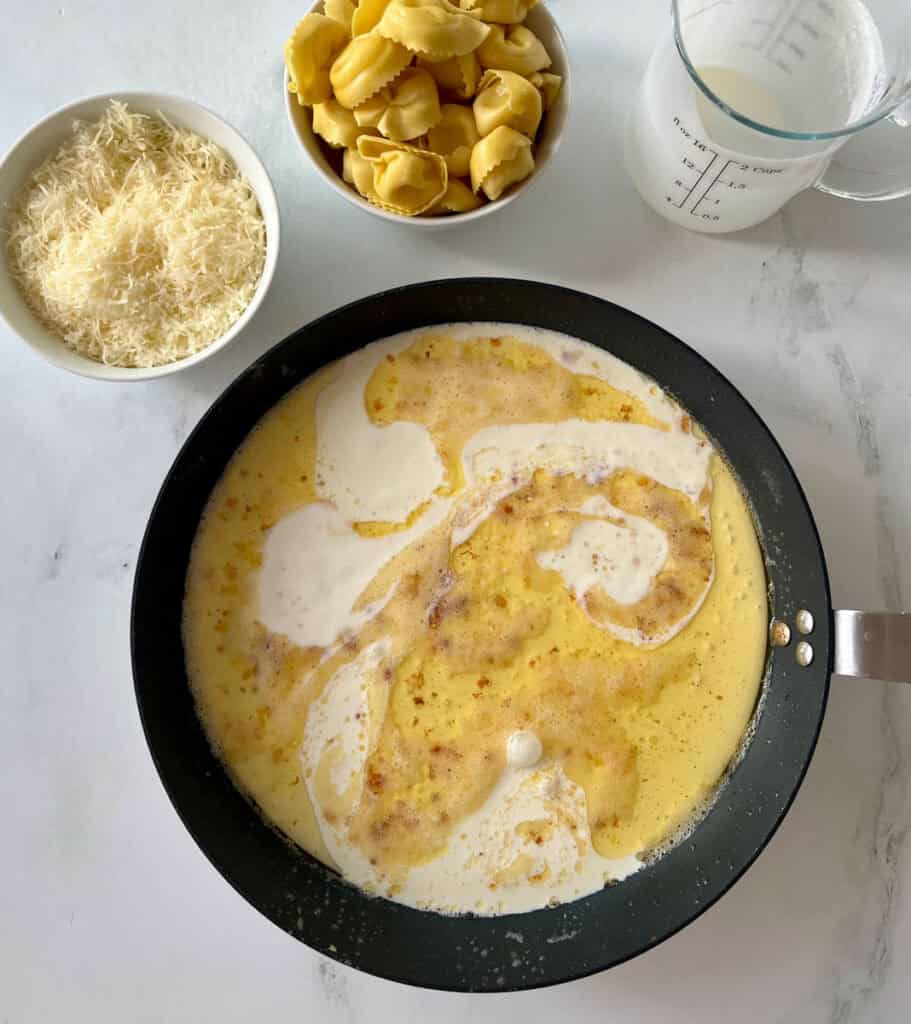A black skillet with a cream sauce with melted butter and seasonings. There is a bowl of uncooked tortellini pasta and grated cheese in the background on a white surface.