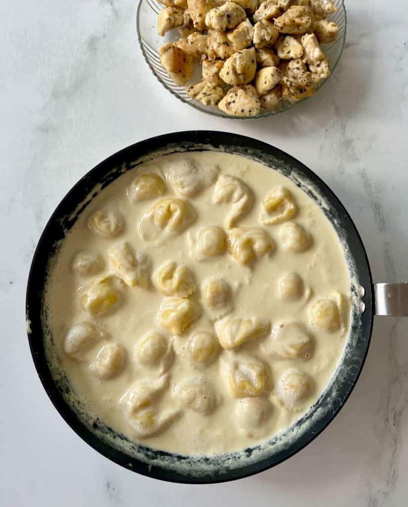 A skillet filled with a creamy sauce and cheese tortellinis alongside a bowl of cooked chicken pieces on a white surface.