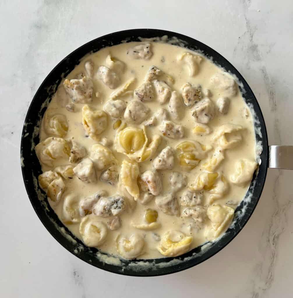 A skillet filled with a creamy sauce, cheese tortellini, and chicken pieces on a white surface.