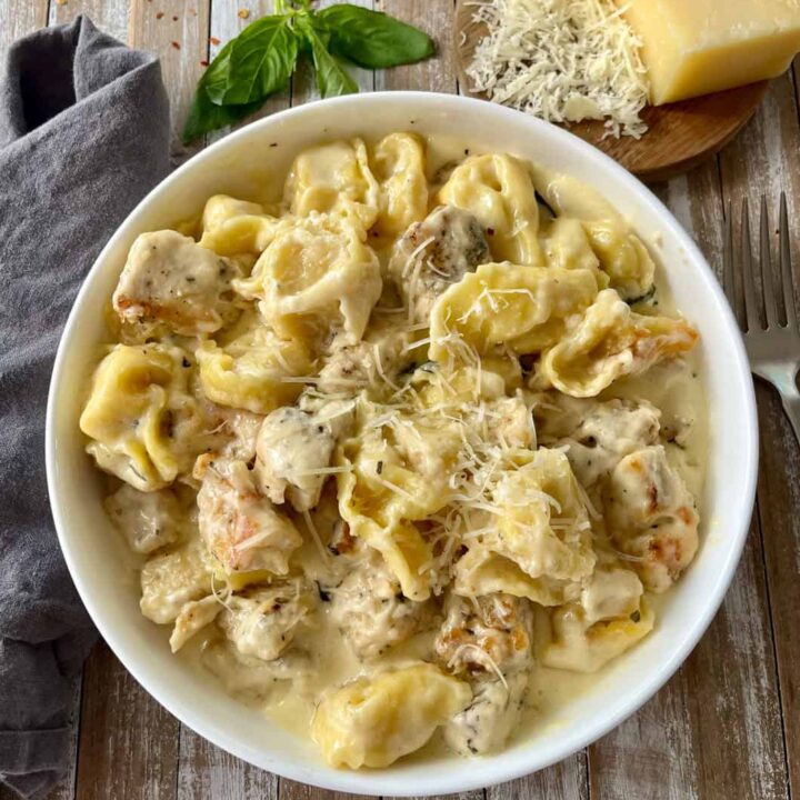 Delicious creamy chicken and tortellini pasta dish with cheese on a rustic wooden surface with cheese, basil, and chili flakes.