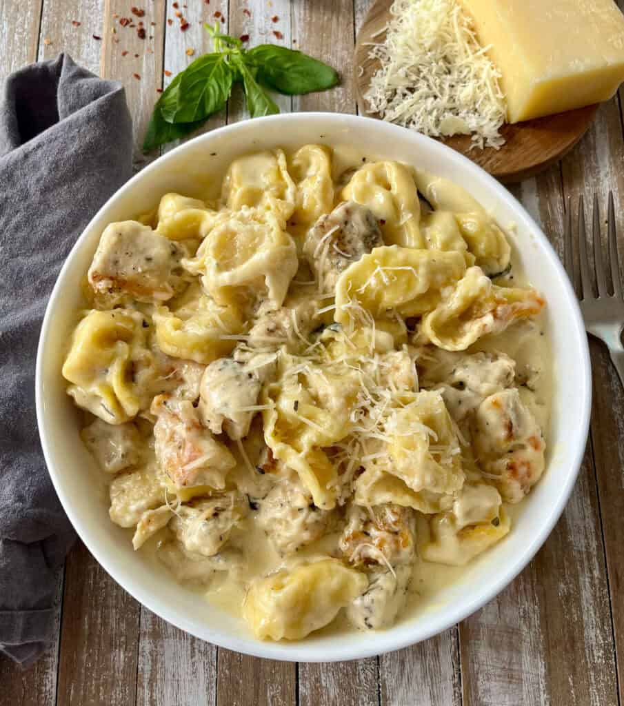 Delicious creamy chicken and tortellini pasta dish with cheese on a rustic wooden surface with cheese, basil, and chili flakes.