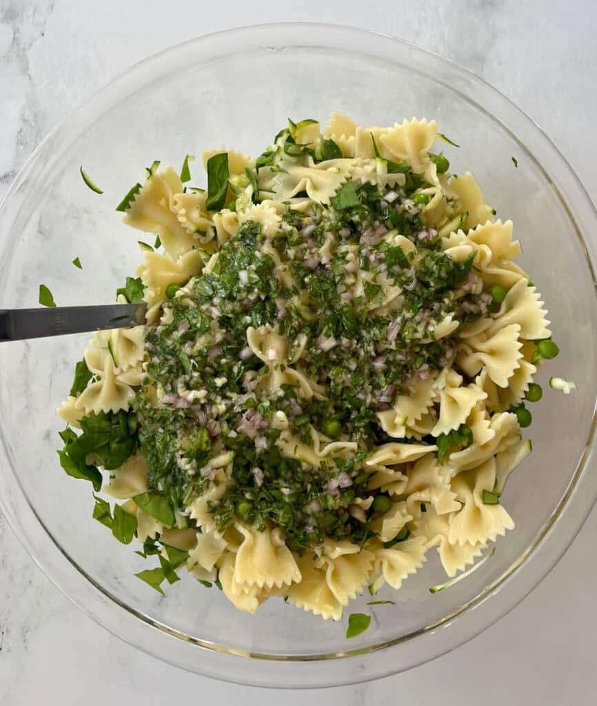 A clear glass bowl filled with cooked bowtie pasta, chopped herbs, and vegetables, garnished with cheese and herbs on a marbled surface.