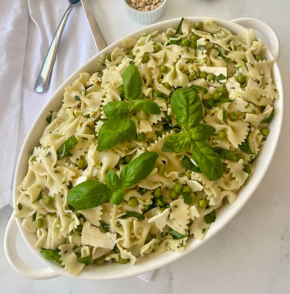 Pasta salad with zucchini, spinach, peas, basil, and lemon basil vinaigrette, garnished with fresh basil and pine nuts on a marbled surface.
