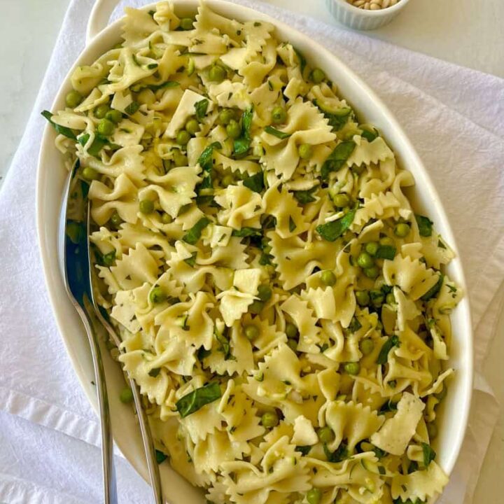 Pasta salad with zucchini, spinach, peas, basil, and lemon basil vinaigrette, garnished with fresh basil and pine nuts on a marbled surface.