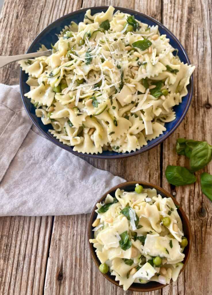 Close-up of cold pasta with shredded zucchini, spinach, peas, basil and cheese in a bowl on a rustic wood surface.