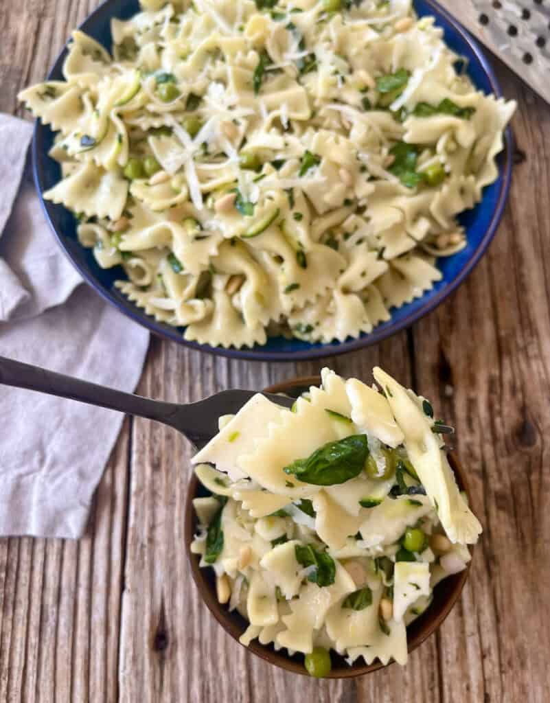 Close-up of cold pasta with shredded zucchini, spinach, peas, basil and cheese in a bowl on a rustic wood surface.