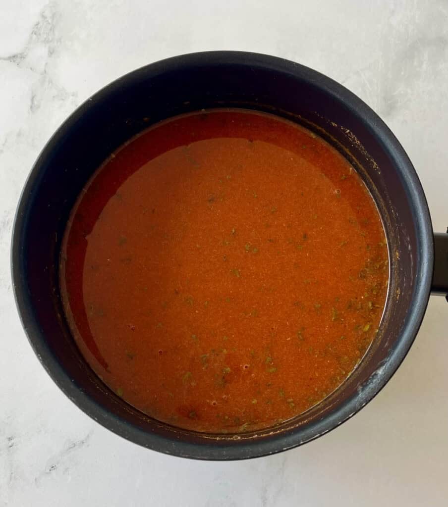 Homemade enchilada sauce in a black saucepan on a marbled surface.