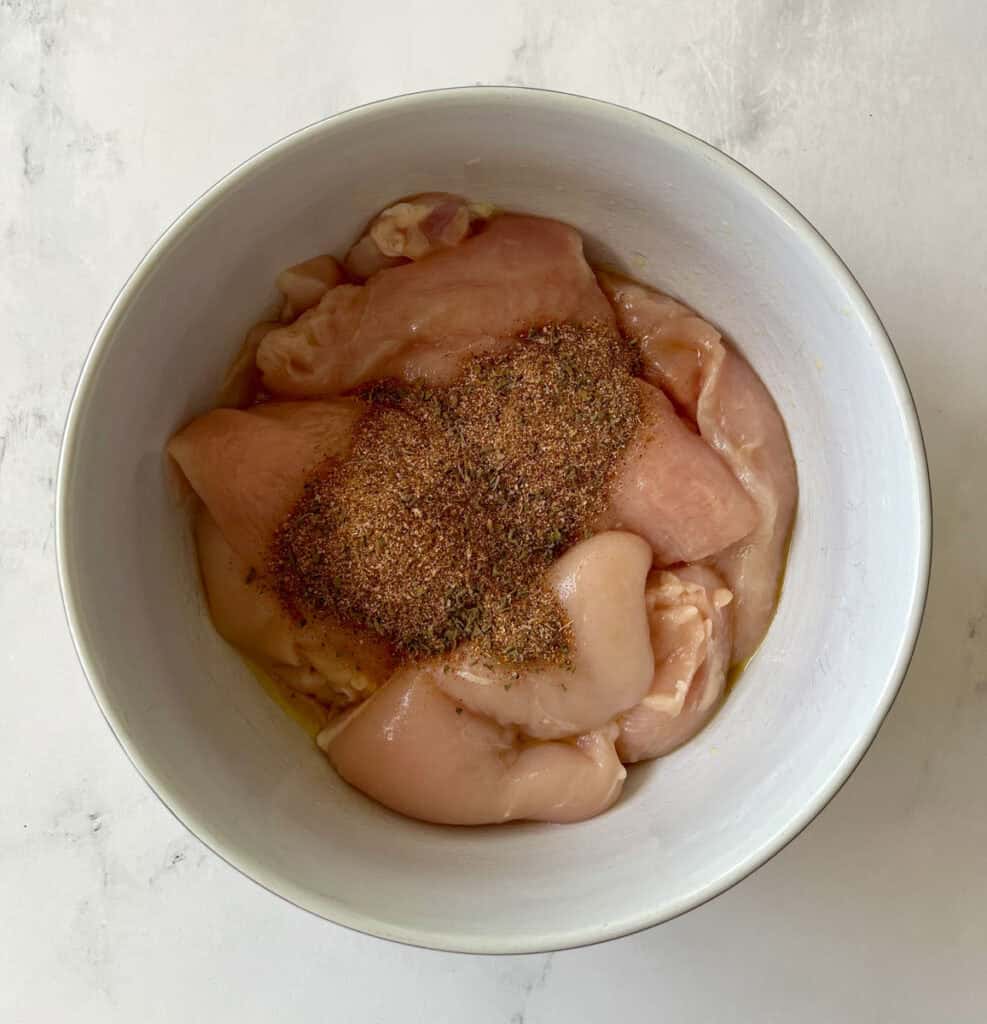 Raw chicken breasts seasoned with spices in a white bowl.
