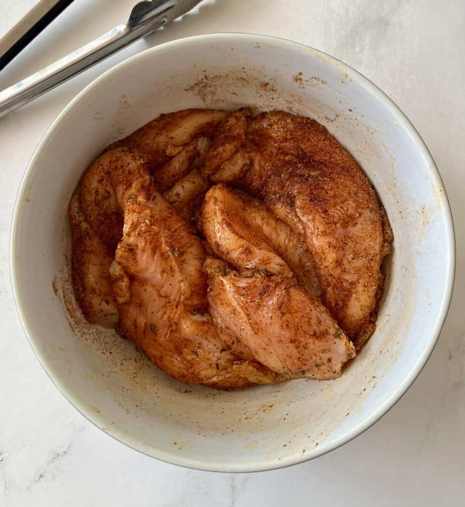 Seasoned chicken breasts in a white bowl, on a marbled surface.