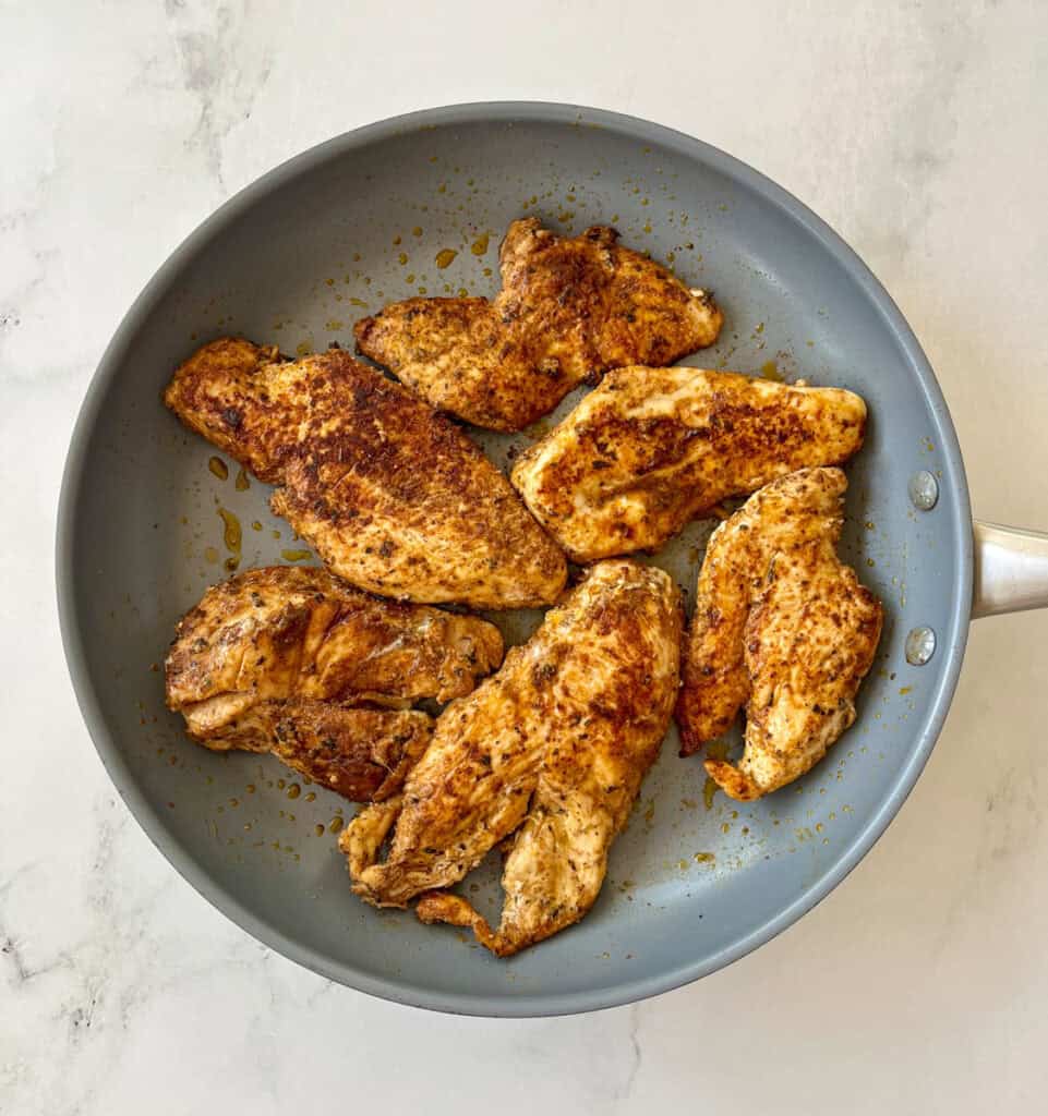 Grilled chicken breasts cooking in grey skillet on a marbled surface.