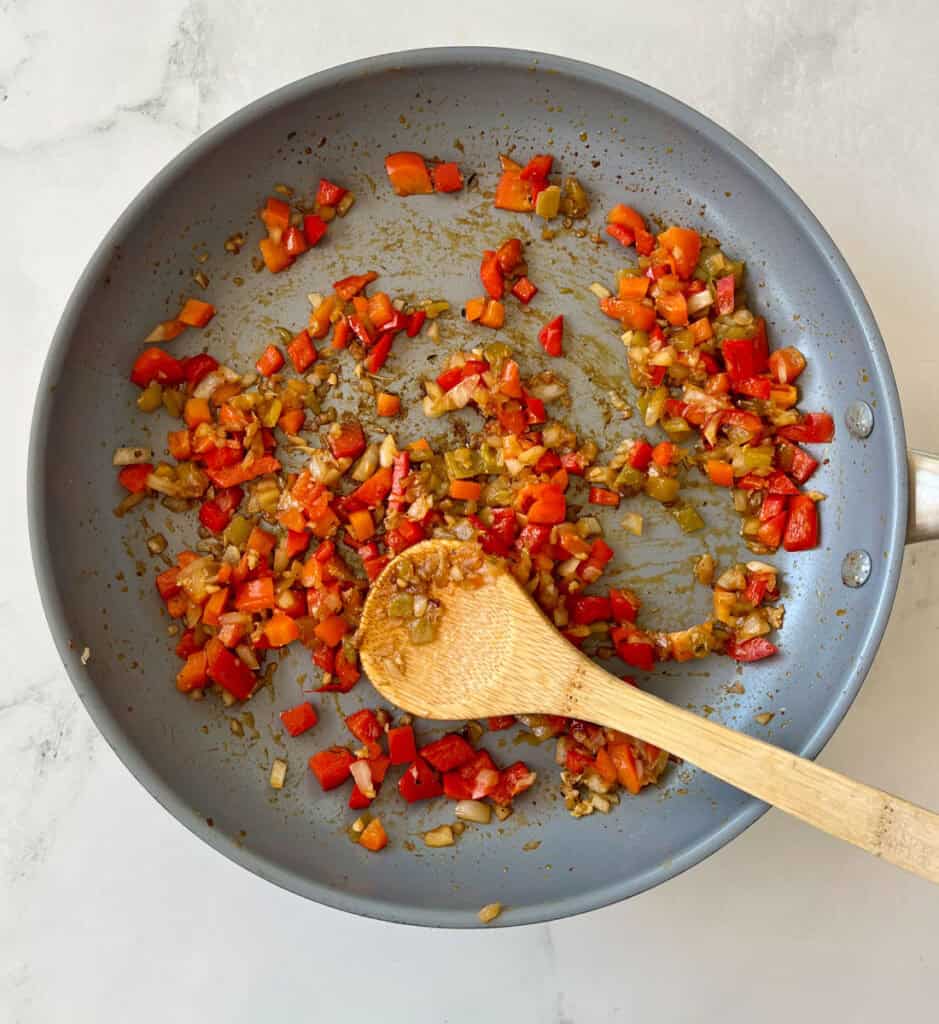 Chopped red bell peppers and onions being sautéed, with a wooden spoon resting inside a grey skillet, on a light surface.