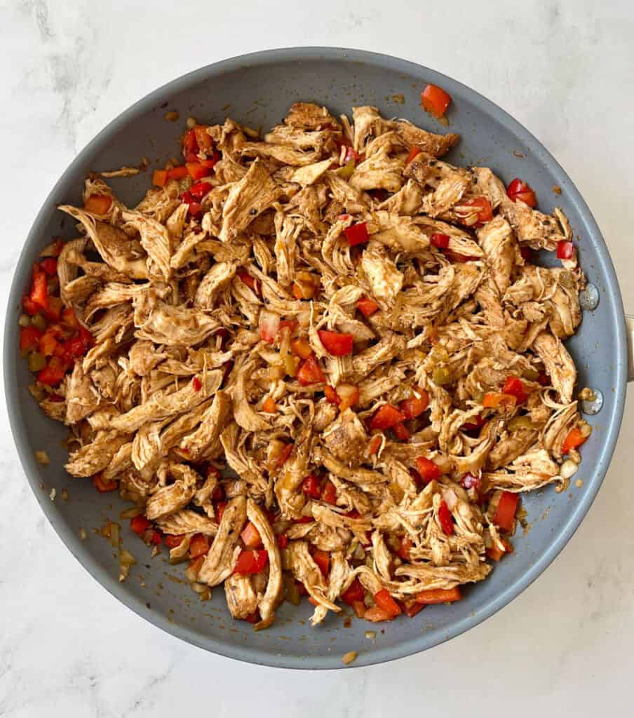 Cooked shredded chicken mixed with diced red bell peppers in a skillet.