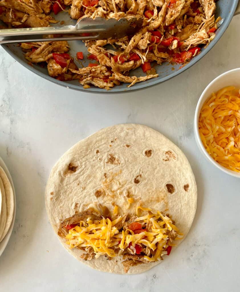 Chicken mixture on a flour tortilla, with a skillet of a chicken mixture on a marbled surface.