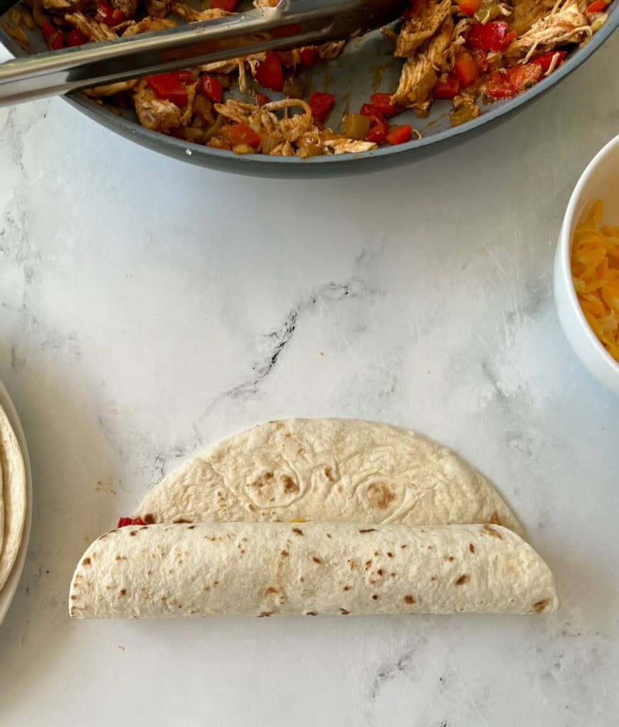 Flour tortilla wrap, with a skillet of a chicken mixture on a marbled surface.