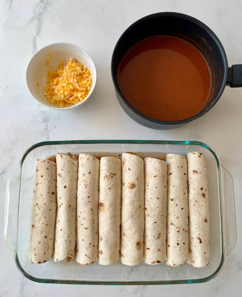 Chicken enchiladas ready to bake, with a pan of sauce and a bowl of shredded cheese on a marbled surface.