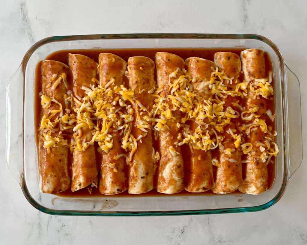 Unbaked homemade chicken enchiladas with sauce and cheese in a clear glass dish on a white surface.