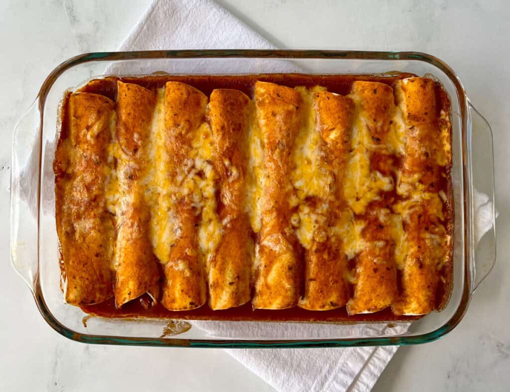 Baked homemade chicken enchiladas with sauce and melted cheese in a clear glass dish on a white surface.