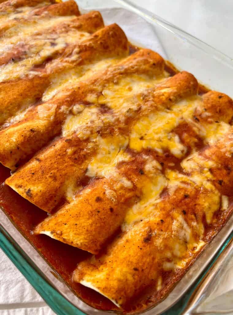 Close-up of homemade chicken enchiladas with sauce, melted cheese and herbs served in a clear glass dish on a white surface.