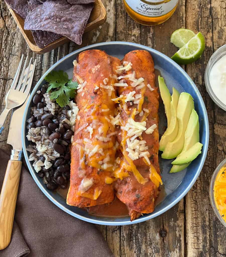 Enchiladas served with fresh avocado slices, black beans, and rice on a rustic wooden table.