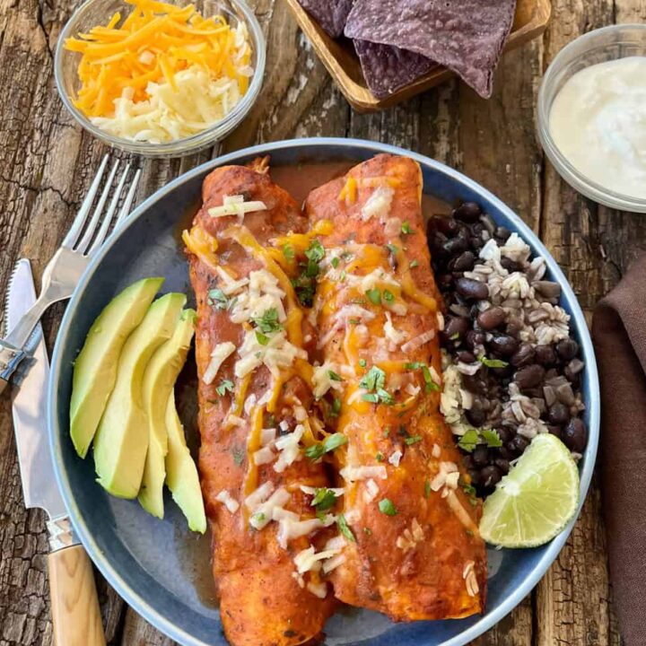 Enchiladas served with fresh avocado slices, black beans, and rice on a rustic wooden table.
