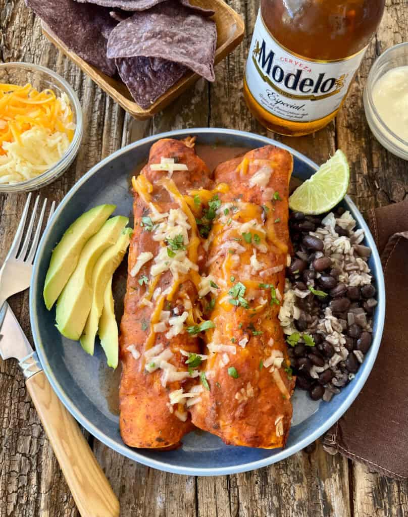 Enchiladas served with fresh avocado slices, black beans, and rice on a rustic wooden table.