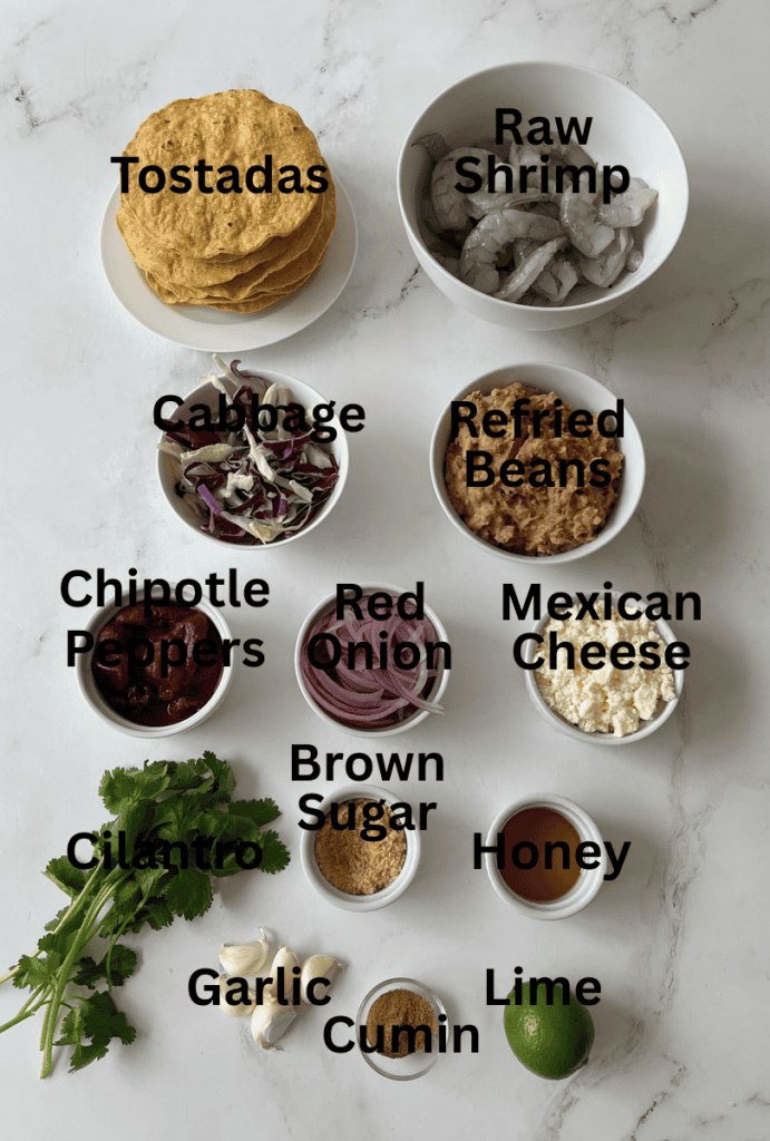 Assorted fresh ingredients for tostadas including shrimp, cabbage, refried beans, fresh herbs, and spices on a marbled surface