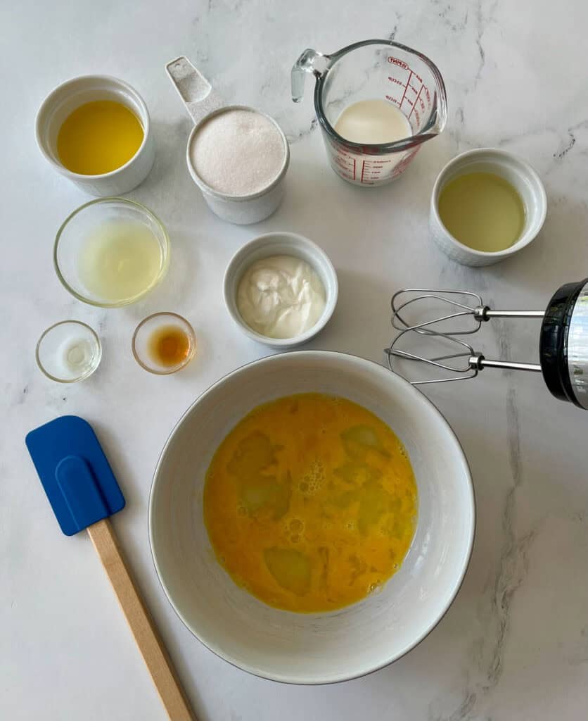 Various baking ingredients like milk, oil, and vanilla extract, along with a hand mixer and a large bowl with lightly beaten eggs.