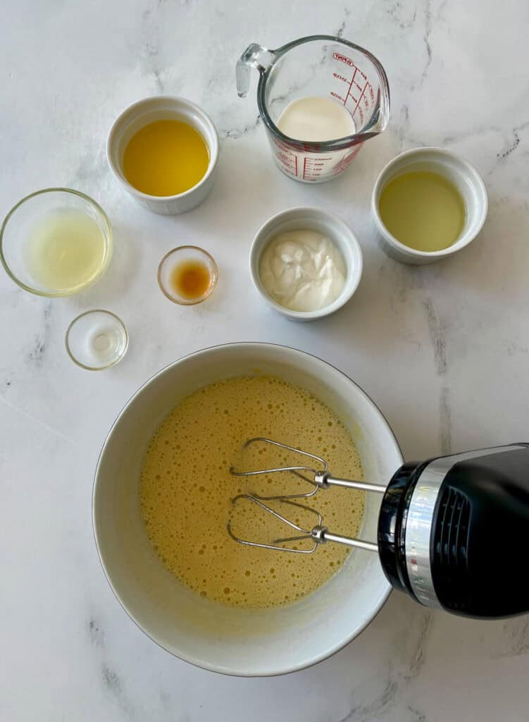 Various baking ingredients like eggs, milk, oil, and vanilla extract, along with a hand mixer blending a yellow batter.