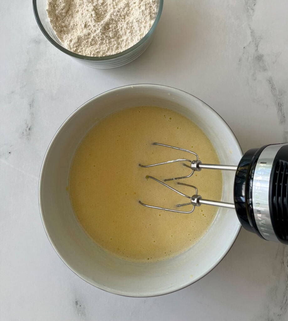 Hand mixer blending yellow batter in a white mixing bowl.