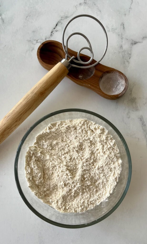 Bowl of flour with wooden measuring spoons and mixing tool on marble surface.