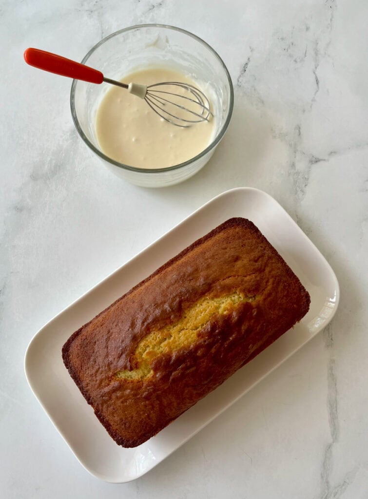 A freshly baked lemon loaf on a white plate accompanied by a bowl of white lemon icing and a whisk.