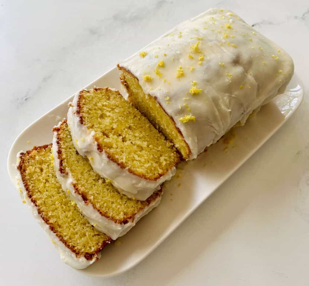 Freshly baked lemon loaf sliced on a white platter, showcasing its moist texture and zesty lemon topping.