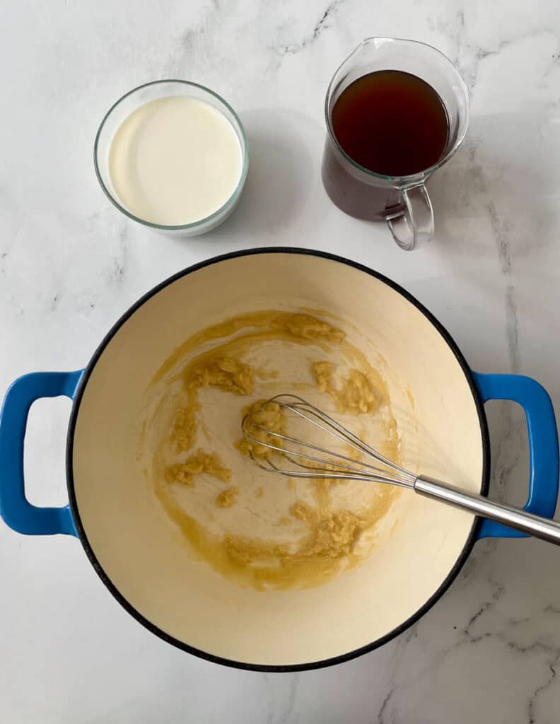 Whisking a butter and flour mixture in a blue pot with milk and broth on a marble countertop.