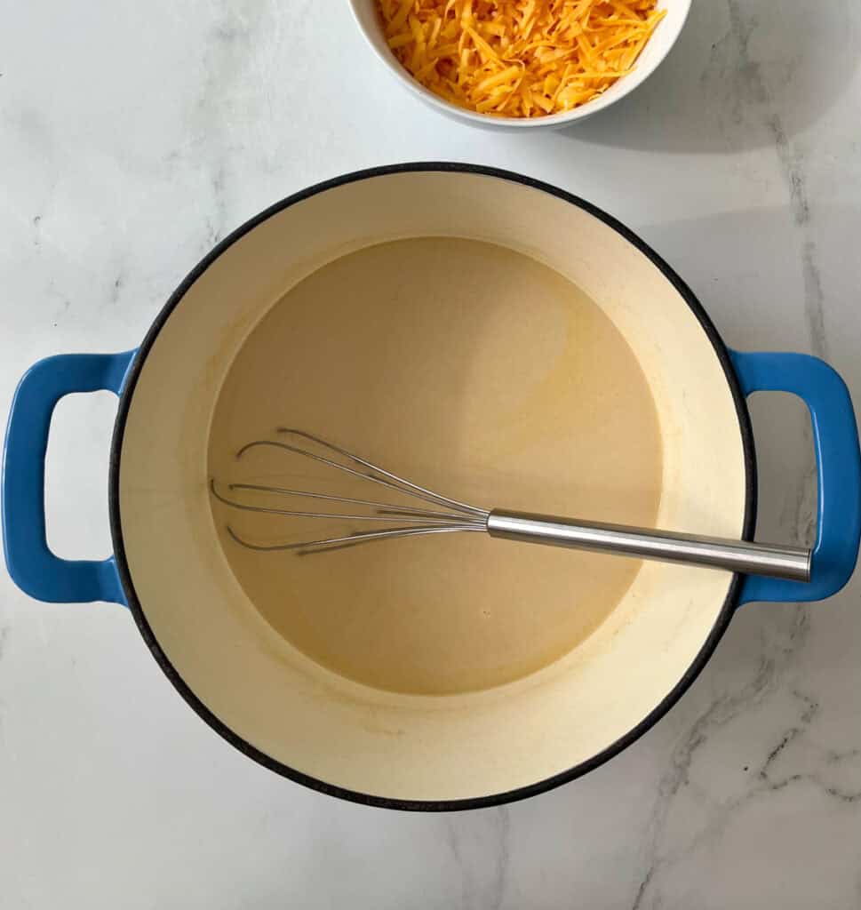 A large pot with sauce and a whisk inside on a white marble countertop, with a bowl of shredded cheese nearby