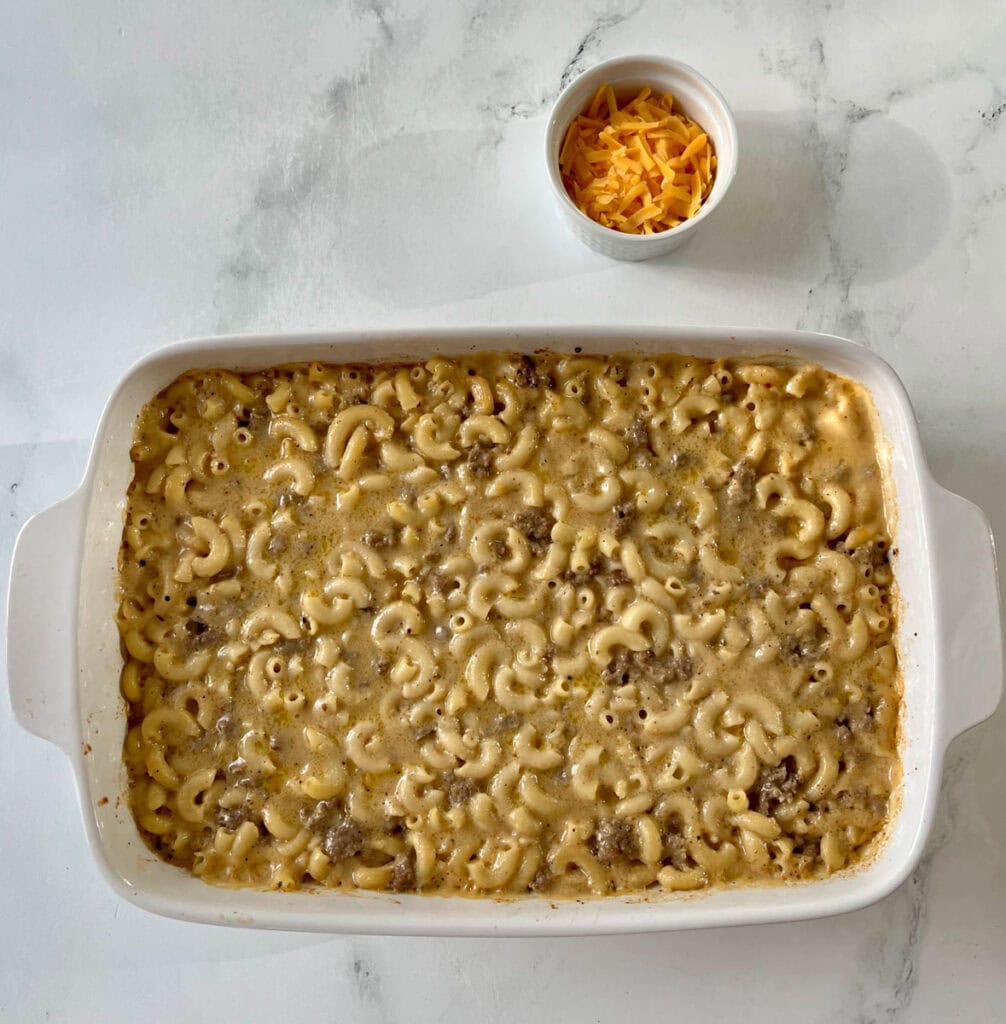 Comforting baked macaroni and cheese with a golden cheese topping in a white baking dish. There is a small bowl of shredded cheese.
