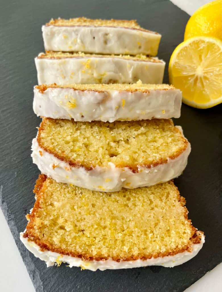 Lemon loaf slices with icing on a black slate board, garnished with lemon halves.