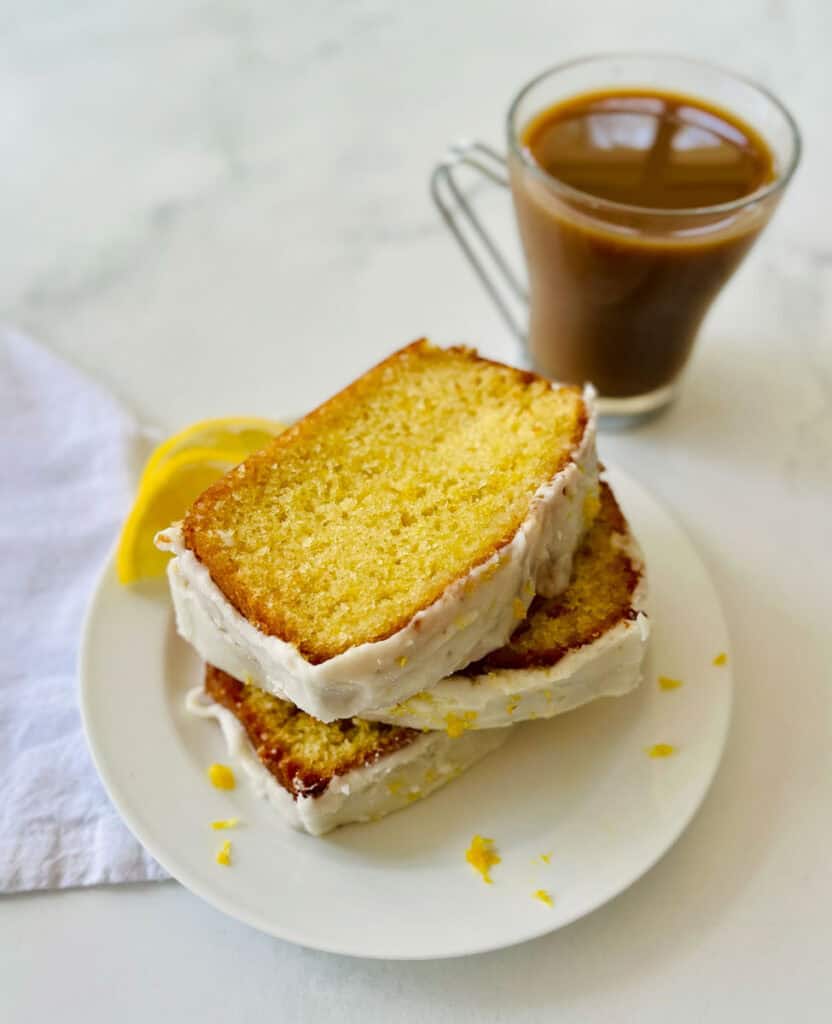 A close-up of sliced lemon loaf with a lemon glaze and lemon wedges on a white plate.