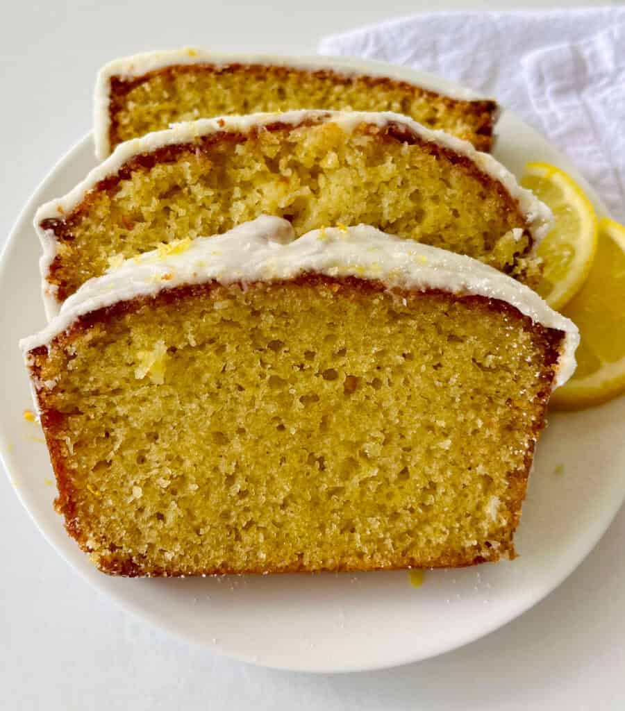 A close-up of sliced lemon loaf with a lemon glaze and lemon wedges on a white plate.
