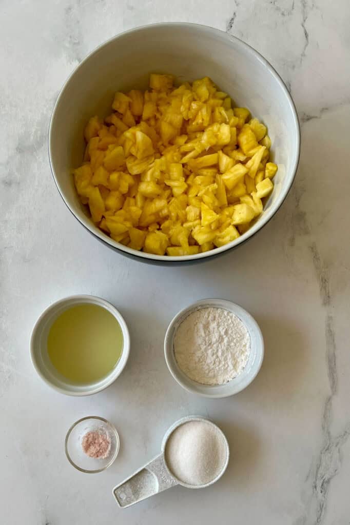 Chopped pineapple pieces in a bowl, with small bowls of lemon juice, cornstarch, sugar, and salt on a marble surface.