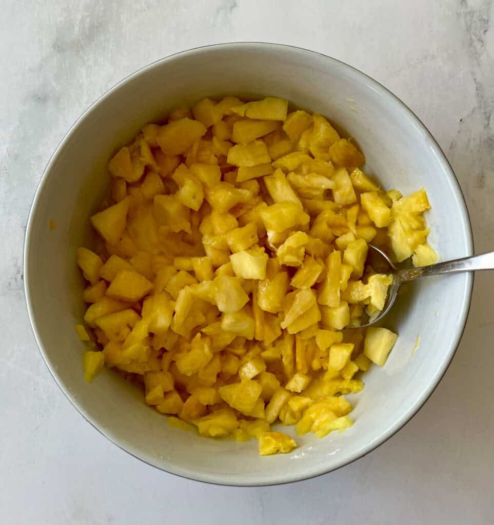 Freshly chopped pineapple pieces with sugar and other ingredients in a white bowl with a spoon.