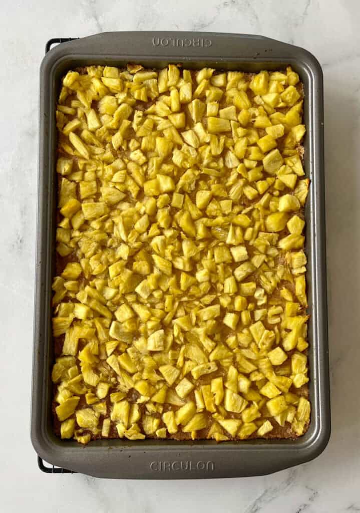 Baked crust topped with golden, diced pineapple pieces in a baking pan, cooling on a wire rack.