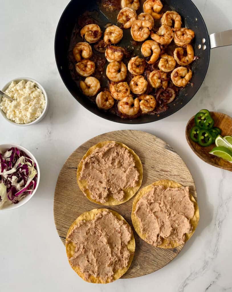 Three crispy tostadas topped with creamy refried beans on a wooden serving board. Spicy shrimp, shredded cabbage, and lime wedges on a white background.