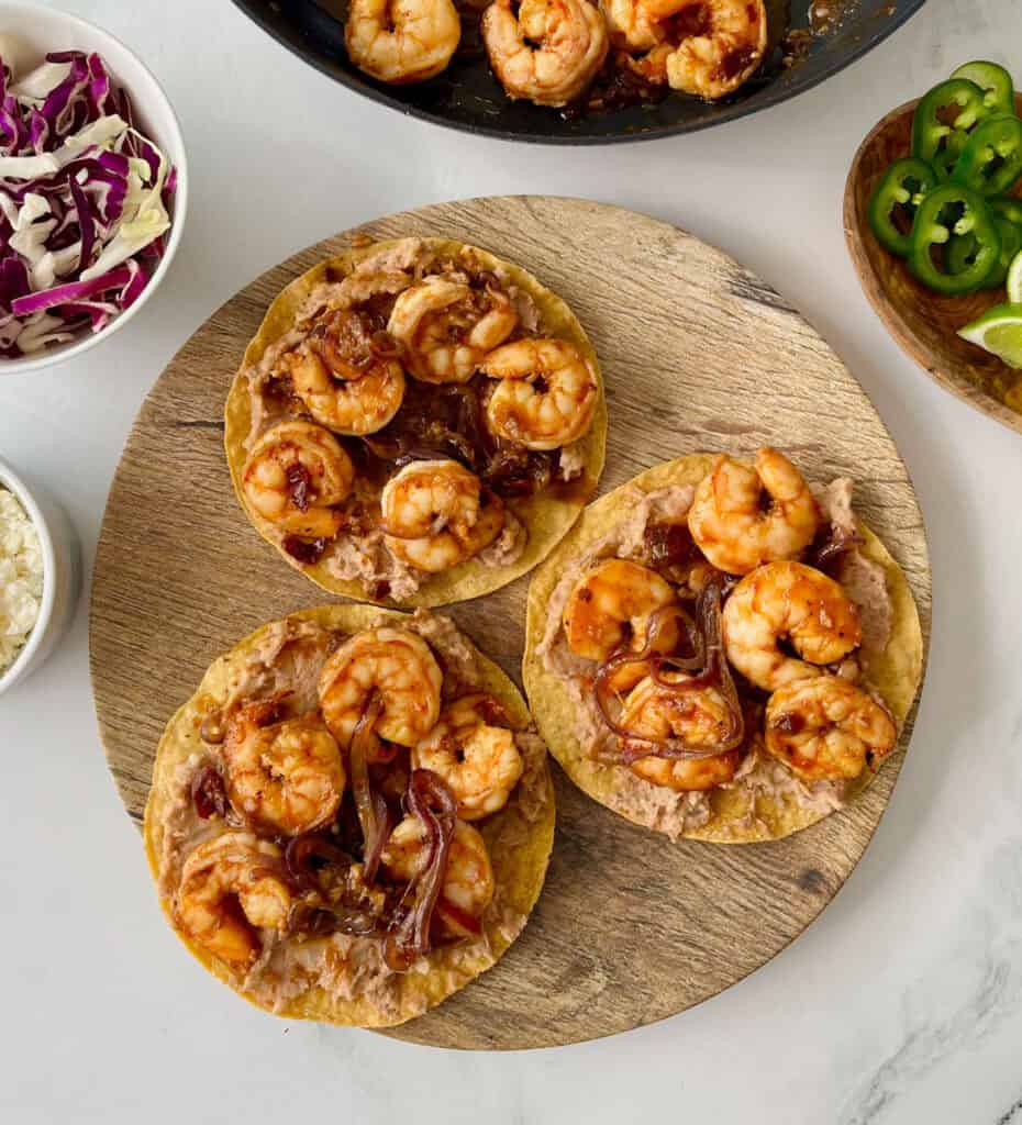 Three shrimp tacos with onions, refried beans, and sauce on a wooden serving board.