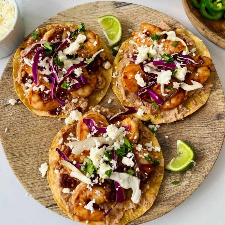 Three shrimp tostadas topped with cheese, onions, and sliced limes on a wooden serving board.