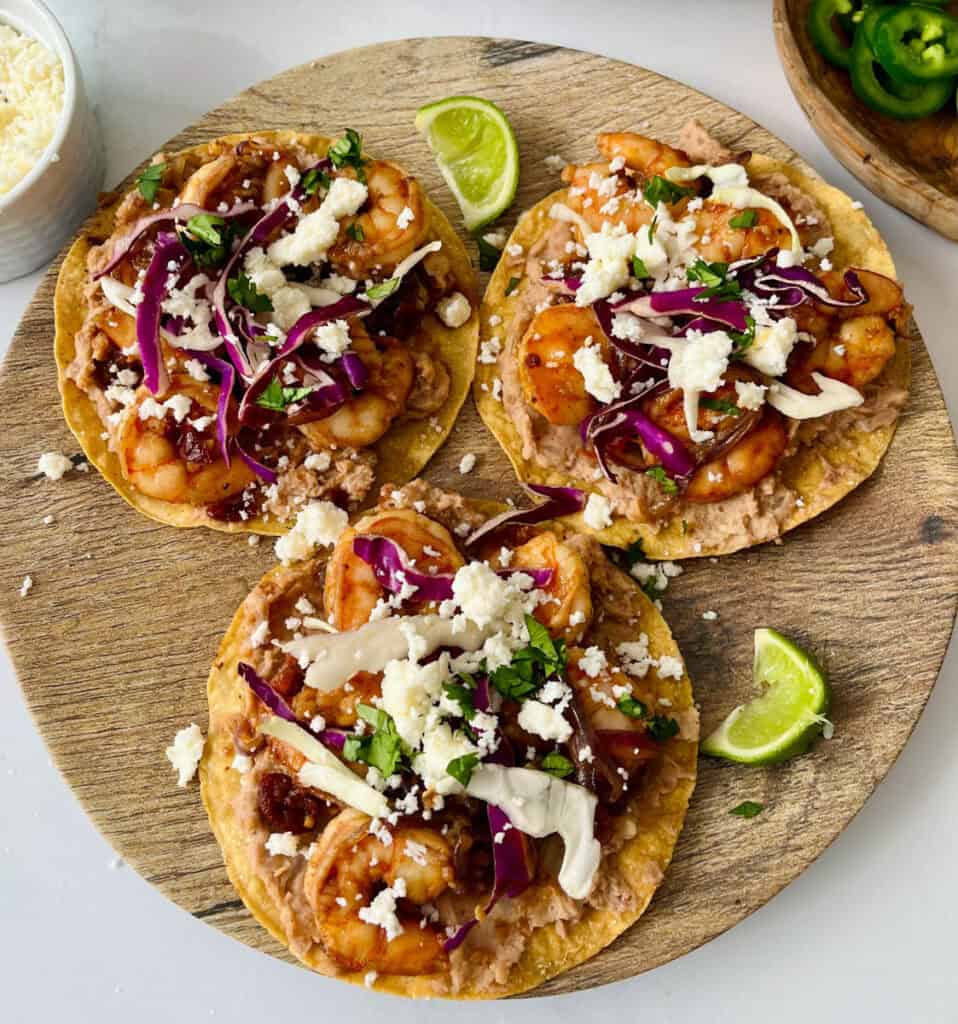 Three shrimp tostadas topped with cheese, onions, and sliced limes on a wooden serving board.