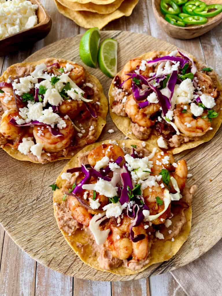 Closeup image of three shrimp tostadas topped with cheese, onions, and sliced limes on a wooden serving board with dishes of crumbled cheese, tostadas, and sliced jalapeño peppers.