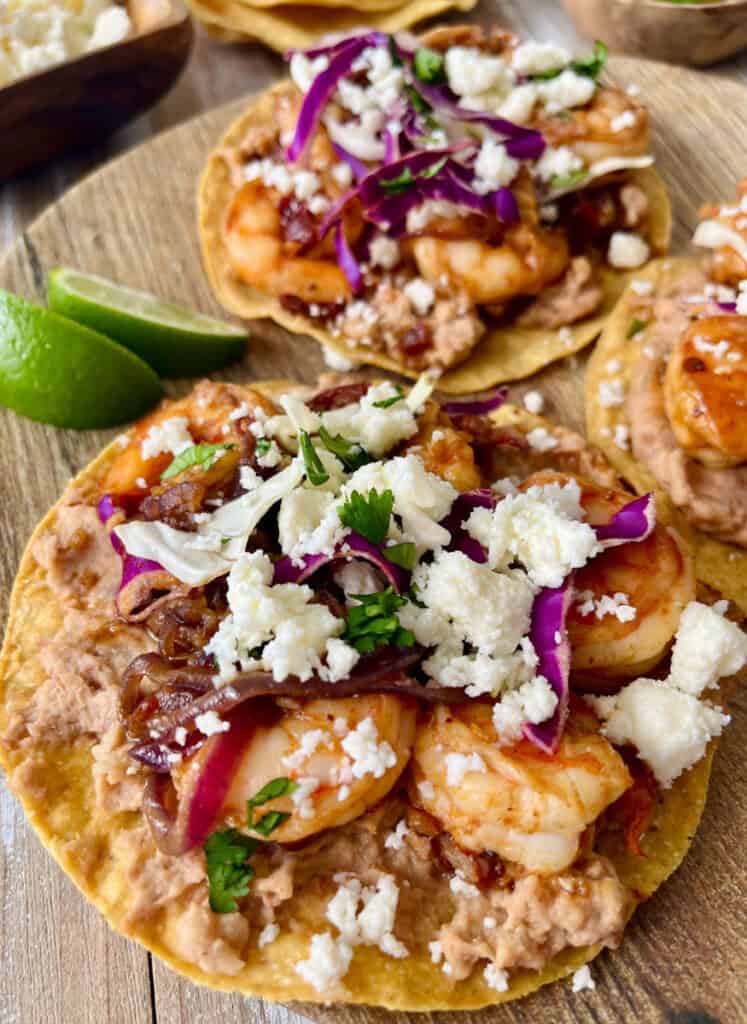 Closeup of shrimp tostadas topped with cheese, onions, and sliced limes on a wooden serving board.